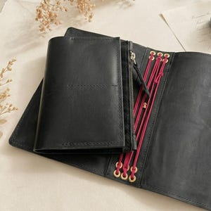 May include: A black leather traveler's notebook cover featuring a zippered pouch insert and a series of vertical magenta elastic cords secured with gold-toned metal eyelets. The cover is open, displaying the interior structure and attachment system.