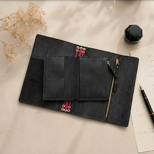 May include: A black leather notebook cover featuring a smaller attached wallet insert, secured with bright pink elastic cords threaded through gold-toned eyelets. The cover includes a vertical gold-toned zipper pocket and multiple interior card slots.