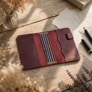 May include: A deep burgundy leather journal cover featuring an interior with multiple elastic bands for securing notebooks, a pen loop, and a side pocket. The name LIA is embossed in gold on the right interior flap, which has a scalloped edge.