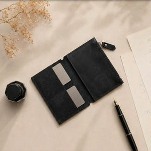 May include: A black leather wallet featuring a zippered compartment and two interior card slots. The wallet is open, revealing a smooth matte texture. A black inkwell and a black fountain pen are positioned nearby on a light surface.