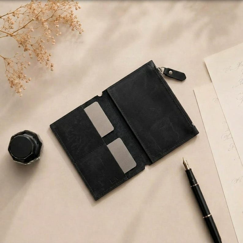 May include: A black leather wallet featuring a zippered compartment and two interior card slots. The wallet is open, revealing a smooth matte texture. A black inkwell and a black fountain pen are positioned nearby on a light surface.