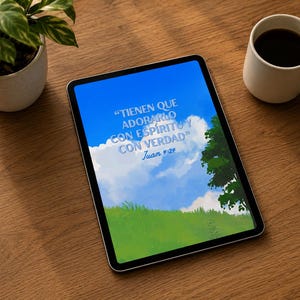 May include: A tablet displaying a digital illustration of a bright blue sky with white clouds, green grass, and a tree. The text reads: "TIENEN QUE ADORARLO CON ESPÍRITU Y CON VERDAD" Juan 4:24. The tablet is positioned on a wooden surface near a plant and a mug.