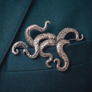 Op de afbeelding: Een zilverkleurige metalen broche in de vorm van een octopus met meerdere krullende tentakels. Het oppervlak van de broche heeft een getextureerde, gepitte afwerking met verhoogde cirkelvormige zuignapdetails langs de lengte van elke arm, vastgespeld op stof.