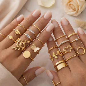 Peut inclure: Une collection de bagues dorées portées sur les doigts, présentant divers motifs dont une rose, un papillon, un cœur, des feuilles, un cercle, une pierre taille baguette, des perles, un anneau torsadé et un large anneau lisse et brillant.