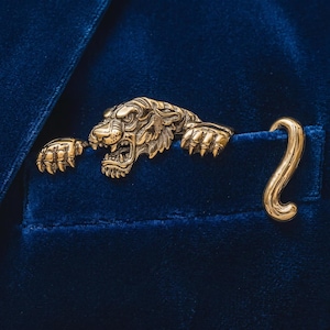 Puede incluir: Un broche de metal dorado con forma de tigre, que presenta una cabeza detallada con dientes al descubierto, dos patas delanteras y una cola curva. El broche está sujeto a una tela de terciopelo azul intenso, creando la apariencia de un tigre asomándose.