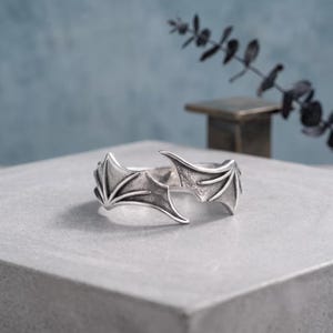Puede incluir: Un anillo abierto de tono plateado que presenta dos diseños simétricos de alas de murciélago que se encuentran en el centro. El metal tiene un acabado oxidado con áreas oscuras hundidas que resaltan los detalles texturizados de las alas.