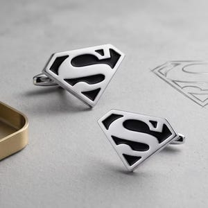 Puede incluir: Un par de gemelos de metal plateado con el icónico logotipo de Superman en forma de diamante con una incrustación de esmalte negro. El diseño muestra la letra S centrada dentro del marco en forma de escudo, con un acabado metálico pulido.