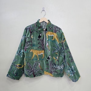 Puede incluir: Una chaqueta de manga larga con cremallera en tejido de terciopelo verde suave con un estampado de temática selvática. El patrón incluye ilustraciones de leopardos, tucanes, loros y cebras entre palmeras y follaje tropical.