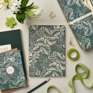 May include: A green and white patterned journal with the words "My Book Journal" on the cover. The journal is surrounded by other stationery items, including a green and white patterned card, a green ribbon, and a green and white patterned gift wrap.