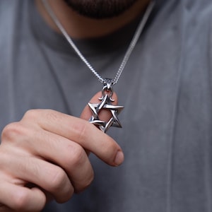 Könnte beinhalten: Ein silberfarbener Davidstern-Anhänger mit polierter Metalloberfläche und einem komplexen, ineinander verschlungenen Design, der an einer passenden silberfarbenen Kette vor einem grauen Stoffhintergrund hängt.
