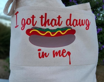 Peut inclure: Un sac fourre-tout en toile beige avec le texte rouge I got that dawg in me au-dessus et en dessous d'un graphique de hot-dog avec une saucisse rouge, de la relish verte et de la moutarde jaune. Le sac a une anse et une forme rectangulaire.
