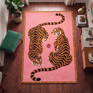 May include: A rectangular area rug featuring a soft pink background with a dark brown border. Two tigers with orange fur and black stripes are depicted on the rug, one lying down and one sitting, with a small decorative symbol between them.