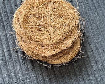 May include: A circular, nest-like structure composed of thin, light brown, fibrous plant material. The object features a tangled, woven appearance with loose, protruding strands creating a textured, layered surface that is round in shape.