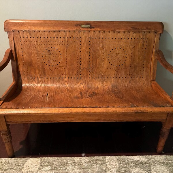 May include: A wooden bench with a curved backrest featuring two rectangular panels of perforated patterns. The bench has curved armrests, four turned legs, and a small metal plaque centered on the top rail of the backrest.