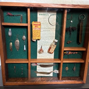 May include: A wooden display case with a glass front containing vintage fishing lures, hooks, and a small bottle of Doodle Oil. The green felt-lined compartments hold various metal spoons, feathered jigs, and a paper advertisement for Doodle Oil.