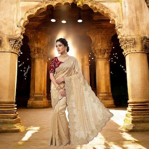 May include: A cream-coloured saree featuring intricate gold embroidery along the scalloped borders, paired with a short-sleeved maroon blouse with gold floral patterns. The garment is draped elegantly, showcasing detailed needlework and a smooth fabric.