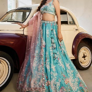 May include: A turquoise lehenga set featuring a sleeveless blouse and a floor-length skirt, both adorned with intricate silver and multi-coloured floral embroidery. A sheer, pale pink dupatta with a decorative border and hanging tassels is draped.