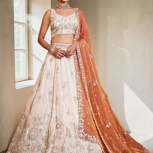 May include: A cream-coloured lehenga set featuring a sleeveless blouse and a floor-length skirt, both adorned with intricate silver and gold floral embroidery. An orange dupatta with a wide, heavily embellished border drapes over one shoulder.