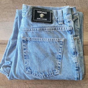 May include: Light blue denim jeans featuring a black rectangular leather patch on the back waistband with the text VERSACE JEANS COUTURE and a silver Medusa head logo. The jeans include a single back pocket with a V-shaped stitched design.