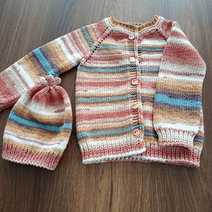 May include: A hand-knitted long-sleeve cardigan and a matching beanie hat. The set features a variegated yarn pattern with horizontal stripes in shades of terracotta, cream, tan, and teal. The cardigan has five small round buttons down the front.