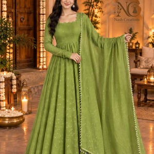 Puede incluir: Un vestido largo de color verde oliva con mangas largas, escote cuadrado y cintura fruncida. La tela presenta un sutil patrón brillante y se combina con un chal dupatta verde oliva a juego que tiene un borde con cuentas blancas.