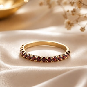 Könnte beinhalten: Ein goldener Ring mit einer durchgehenden Reihe kleiner, runder, tiefroter Granat-Edelsteine in Krappenfassung. Der Ring hat eine polierte metallische Oberfläche und ein schmales, kreisförmiges Profil mit einer gleichmäßigen Anordnung.
