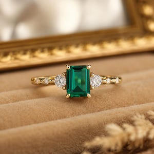 Peut inclure: Une bague en or avec une pierre précieuse verte taille émeraude au centre, flanquée de deux pierres blanches rondes. L'anneau est fin et métallique, orné de petites pierres blanches rondes sur les côtés, formant un design symétrique.