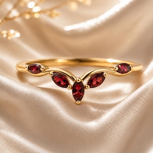 Könnte beinhalten: Ein V-förmiger Goldring mit fünf tiefroten Granat-Edelsteinen im Marquise-Schliff, die in einem symmetrischen Muster entlang der Ringschiene angeordnet sind. Das Metallband ist poliert und die Steine sind in einzelnen Goldfassungen gefasst.
