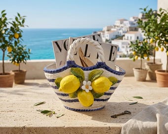 May include: A ceramic basket with a white and blue striped pattern, featuring a sculpted cluster of three yellow lemons and a small white flower on the front. The basket has a braided white handle and contains two magazines labelled Vogue.