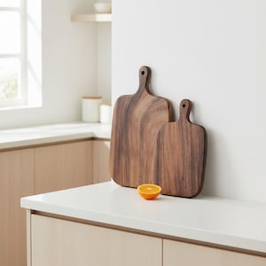 May include: Two dark brown wooden cutting boards with rounded corners and handles, positioned upright. The boards feature a visible natural wood grain pattern. A single halved orange sits on the white surface in front of the boards.