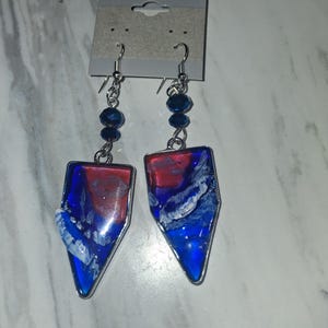 May include: A pair of dangle earrings featuring silver-toned metal frames with a triangular, shield-like shape. The centers contain a marbled resin design in shades of deep blue, bright red, and white. Each earring hangs from a silver hook with a dark blue bead.