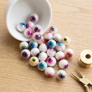 Puede incluir: Una colección de cuentas esféricas con diseños realistas de ojos humanos en varios colores de iris, incluyendo azul, morado, rosa y amarillo, que se derraman de un pequeño cuenco de cerámica blanca, junto a un carrete de latón y alicates.
