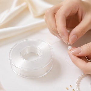 May include: A clear, circular spool of thin, transparent elastic cord used for jewellery making. The cord is being threaded through a small, round, white pearl bead, with a partial strand of white pearls and small gold beads visible nearby.