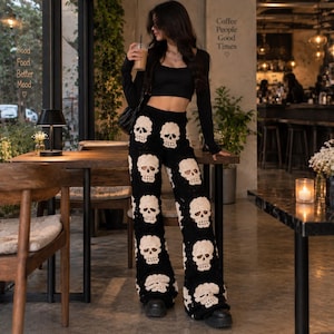 May include: Black long-sleeved crop top paired with high-waisted crochet pants featuring a repeating pattern of cream-colored skulls. The pants have a wide-leg silhouette and are constructed from a black yarn base with detailed skull motifs.
