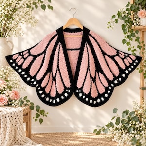 May include: A crochet cardigan designed to resemble butterfly wings, featuring a light pink and black color scheme with small white accents along the scalloped edges. The garment is displayed on a wooden hanger with an open front design.