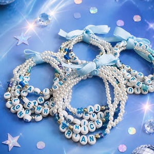 May include: A collection of beaded bracelets featuring white faux pearls, small blue and silver accent beads, and white circular letter beads spelling names like Brian, Nick, and AJ. Each bracelet is tied with a light blue grosgrain ribbon bow.