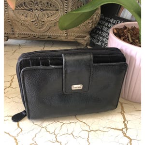 May include: A black leather bi-fold wallet with a wrap-around zip closure and a snap-button flap featuring a small silver-toned metal plaque embossed with the brand name Fossil. The wallet has a textured surface and visible card slots.