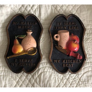 Puede incluir: Dos placas decorativas de pared con fondos negros texturizados y bordes de color bronce. La placa izquierda presenta una pera, una botella y una cuchara con el texto NO MATTER WHERE I SERVE MY GUESTS. La placa derecha muestra una taza, manzanas y un cuchillo con el texto IT SEEMS THEY LIKE MY KITCHEN BEST.