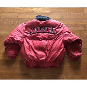 May include: A red puffer jacket featuring a black collar and the word ALABAMA embroidered in red letters with a black and white outline across the upper back. The jacket has long sleeves with elastic cuffs and a ribbed hem at the bottom edge.