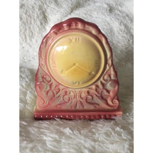 Peut inclure: Une horloge de cheminée en céramique avec un cadran circulaire jaune pâle orné de chiffres romains en relief XII, III, VI et IX. L'horloge possède deux aiguilles et est entourée d'un cadre décoratif aux nuances dégradées rose et crème.