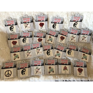 May include: A collection of twenty-two individual removable tattoo sheets from Tattoo Factory, each featuring a unique design such as red roses, yin-yang symbols, peace signs, and birds, all packaged in white cards with black text and graphics.