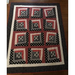 May include: A rectangular quilt featuring a log cabin pattern with twelve blocks. Each block contains a central square of black and white dog print fabric surrounded by red and white polka dot and black and white striped fabric borders.
