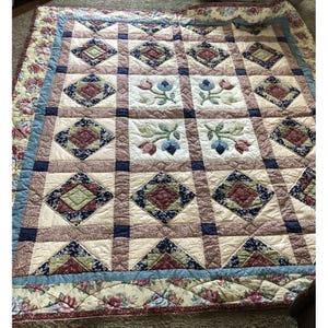 May include: A rectangular quilt featuring a patchwork design of diamond-shaped blocks in shades of burgundy, navy blue, and sage green, interspersed with white squares displaying embroidered floral motifs of blue and pink tulips.