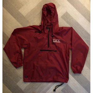 May include: A maroon long-sleeve pullover windbreaker jacket with a hood, featuring a half-zip front closure, a horizontal zippered pocket on the chest, and a drawstring hem. The text U of A is printed in white on the upper left chest area.