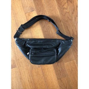 Puede incluir: Una riñonera de cuero negro con un compartimento principal con cremallera, un bolsillo frontal más pequeño con cremallera y una correa ajustable de nailon negro con una hebilla de plástico. El bolso tiene una textura lisa y varios tiradores.