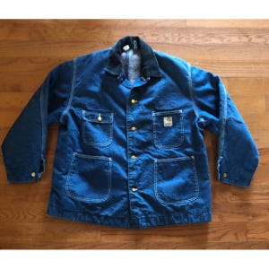 May include: A blue denim chore coat featuring a dark corduroy collar, long sleeves with button cuffs, and a front button closure. The jacket includes two chest pockets, one with a Carhartt label, and two large lower patch pockets.