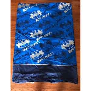 May include: A blue bed sheet featuring a repeating pattern of silver and dark blue Batman logos and the word Batman in a stylized font. The bottom section of the sheet is a solid dark blue color separated by a thin horizontal silver line.