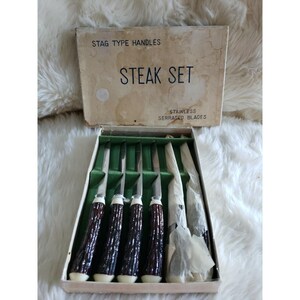 May include: A steak set in a rectangular box with the text STAG TYPE HANDLES and STAINLESS SERRATED BLADES. The set includes four knives with dark brown textured handles and silver blades, plus two additional knives wrapped in white paper.