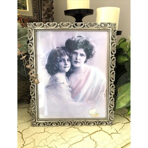 May include: A rectangular silver-toned picture frame with an ornate, embossed rose pattern along the border. The frame contains a vintage-style portrait of two individuals, one child and one adult, with a small price tag visible in the bottom corner.