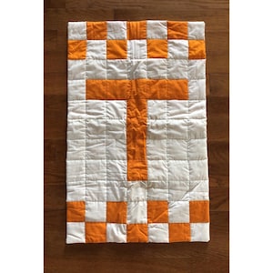 May include: A rectangular quilt featuring a grid of white and orange fabric squares. The center of the quilt displays a large letter T formed by orange squares against a background of white squares, with a border of alternating orange and white squares.